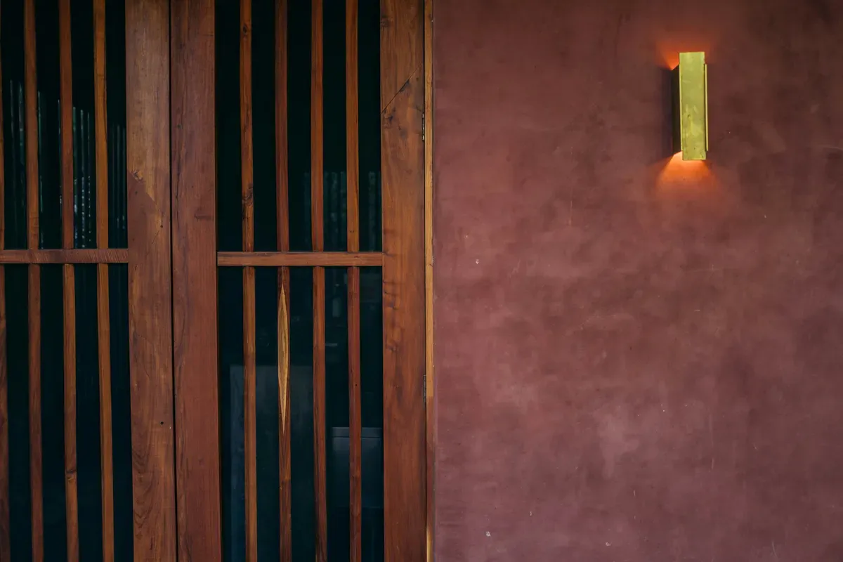 A wooden door set in a red wall with a lamp burning above it