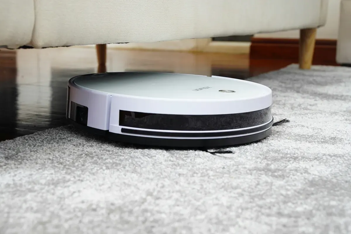 A round robot vacuum crossing from hard floor onto a pale rug beside a sofa